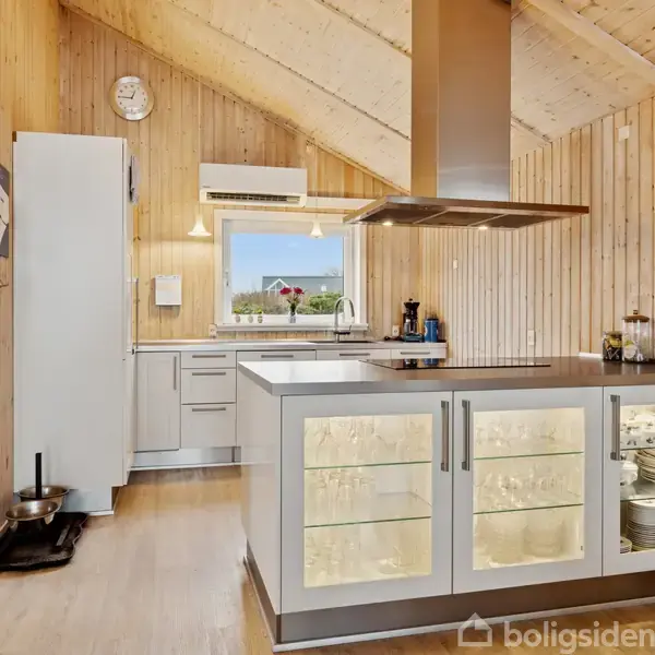 Et lyst køkken med flere skabe, køkkenø og emhætte. Køleskab og opvaskemaskine langs væggen. Trævægge og loft med hældning. Udstyret med kaffemaskine og vindue med udsigt.