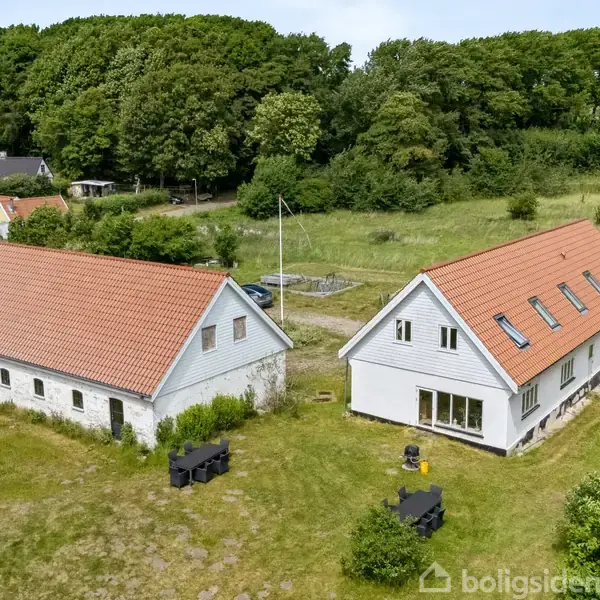 To bygninger med røde tegltage står på en græsklædt grund omgivet af træer. Den ene bygning har store vinduer og flere kviste på taget.