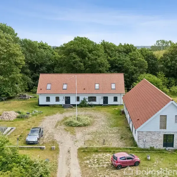 Hvid bygning med rødt tegltag står stille på en åben grund omgivet af træer. To biler holder parkeret på en grusvej foran bygningen.