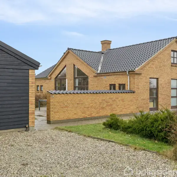 Gulstenshus med sort tag og store vinduer står ved siden af et sort træskur. Grusvej og græsplæne omgiver bygningerne under en blå himmel.