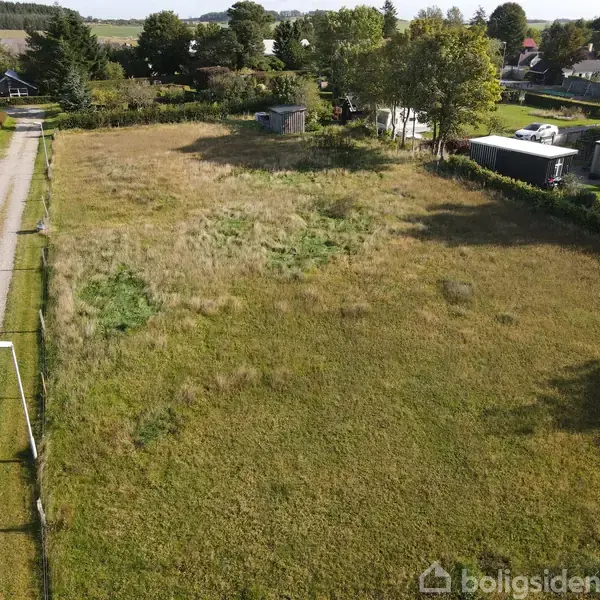 En græsmark med ujævn beplantning omgivet af huse og træer. Til venstre ses en grusvej. I baggrunden er der flere beboelseshuse og en skurbygning.