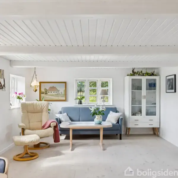 Stue med en blå sofa og et hvidt sofabord centralt placeret. En beige lænestol står til venstre, omgivet af vinduer med blomster. Et skab med glasdør står til højre.