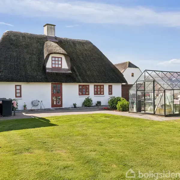 Et stråtækt hus med hvide vægge og røde vinduer står i en solrig have. Ved siden af er et drivhus. Grønne græsplæner omgiver bygningerne under en blå himmel.