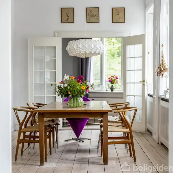 Et spisebord med blomsterbuketter på står i et lyst rum med trægulv. Otte træstole omgiver bordet. Store vinduer og en glasdør åbner rummet ud mod en grøn udsigt.