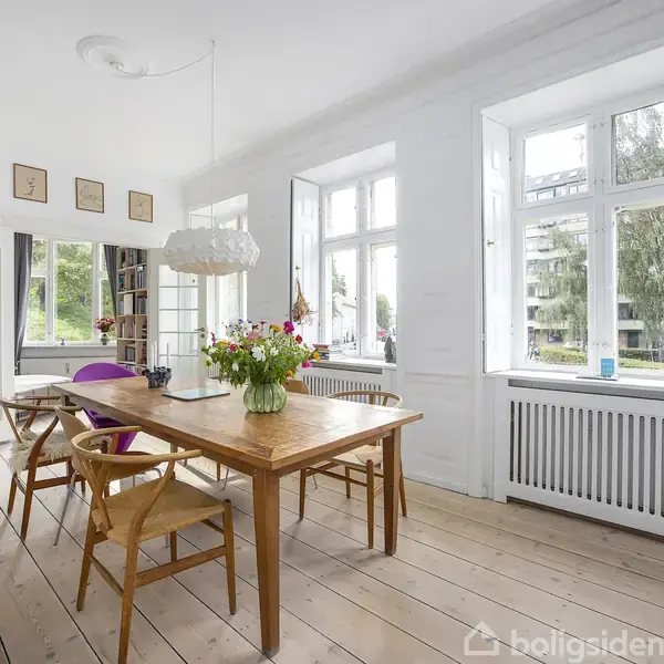 Et stort træbord med blomster og stole står i et lyst spisestueområde. Rummet har trægulve, hvide vægge, store vinduer med udsigt til grønne træer udenfor.
