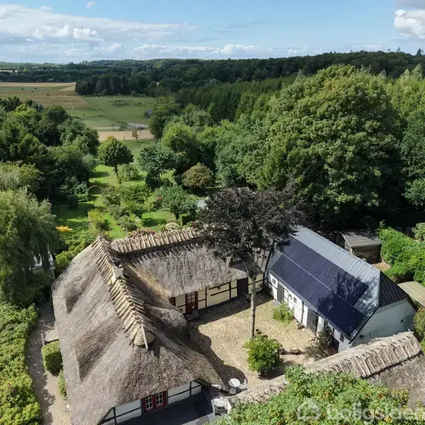 Traditionelt stråtækt bindingsværkshus omgivet af frodig have og skov. Solpaneler er monteret på et af tagene. I horisonten ses åbne marker under en skyfuld himmel.