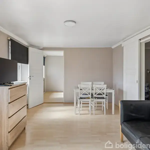 Et værelse har en sort sofa og en kommode med TV. I baggrunden ses et spisebord med hvide stole. Rummet har lyse trægulve og neutrale vægge.