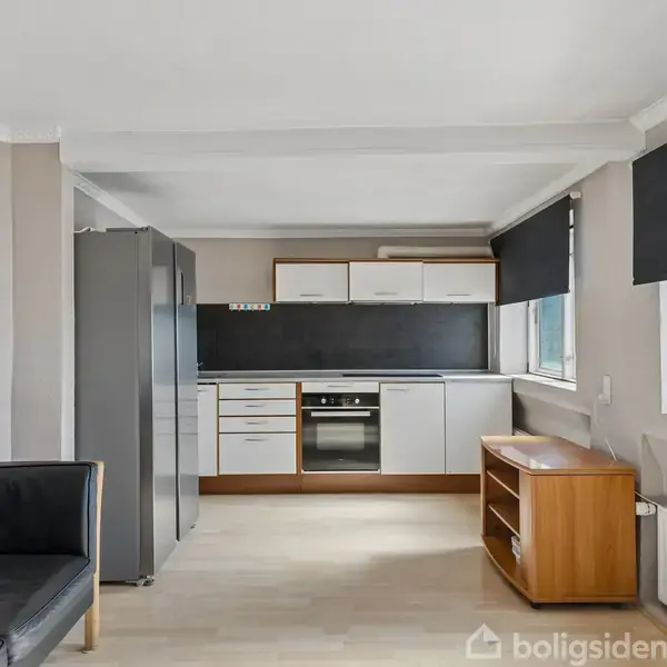 En moderne køkken-alrum viser et køkken med hvide skabe og mørkegrå bordplader. En sort sofa står mod væggen, mens et træskab er placeret mellem to vinduer.