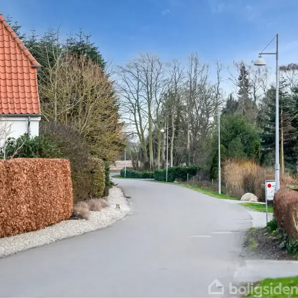 Vejen snor sig forbi et hus med rødt tegltag og hække, i en stille, beboet landskab. Træer står langs vejen, og en lygtepæl er synlig. Nummeret "2" ses på et skilt.