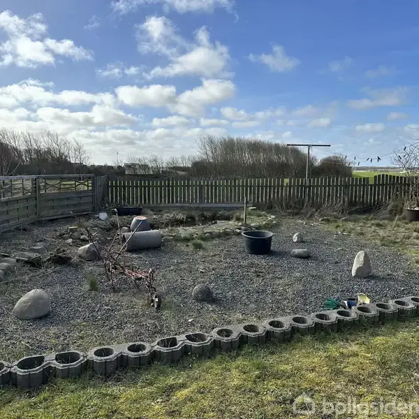 En lille have omgivet af et lavt hegn med grusbelagt jord og spredte sten. Et par plantepotter står tilfældigt placeret. I baggrunden ses åbne marker under en skyet himmel.