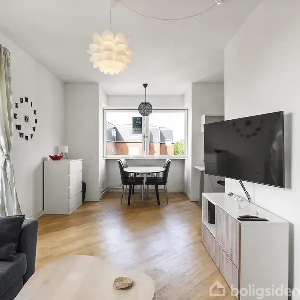 En stue med en fladskærms-TV på væggen, en sofa til venstre og et spisebord ved vinduet. Rum med trægulv og en rund lampe hængende fra loftet.