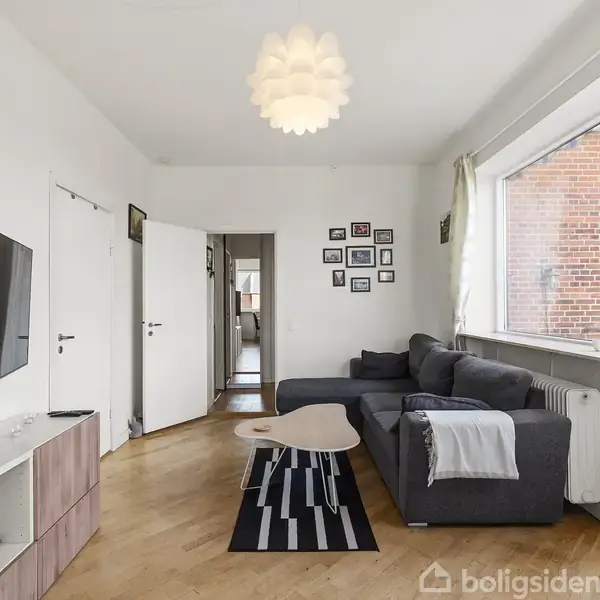 En stue med en grå sofa og et sofabord består centrum. Et tv er vægmonteret ved siden af en lysegrå reol. Et stort vindue med gardiner lader lys ind fra højre.