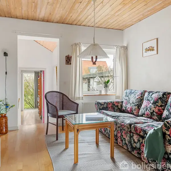 Blomstrede sofa med et bord foran, står i en hyggelig stue. Værelset har hvidmalede vægge, trægulv, og loft, og er dekoreret med billeder og en lampe; vindue slipper lys ind.