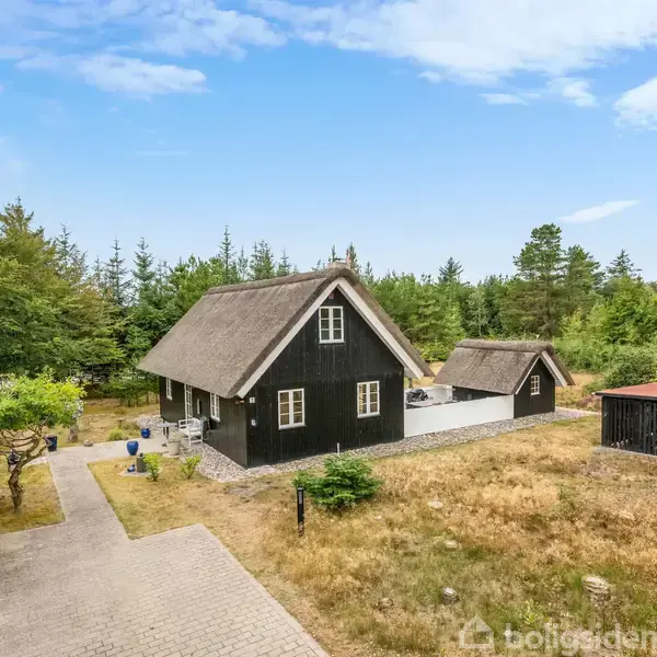 Sort stråtækt hus med en tilstødende lille bygning og skur. Det står på en flisebelagt sti omgivet af græs og træer i naturskønne omgivelser under en blå himmel.