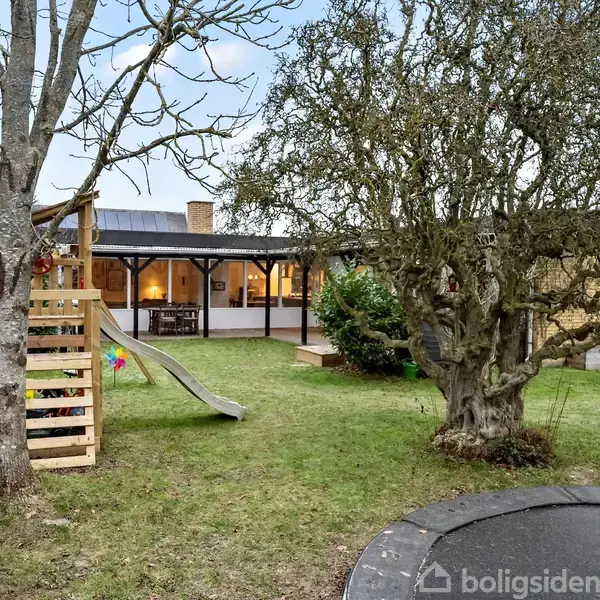 En legeplads med et klatrestativ og rutsjebane står i en have foran et hus. Træer og buske omgiver området, og der er en trampolin i græsset.