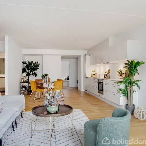 Stue med grå sofa, rundt sofabord, og grøn lænestol i moderne lejlighed. Åbent køkken med hvide skabe og spiseplads med gule stole. Planter tilføjer liv til rummet.