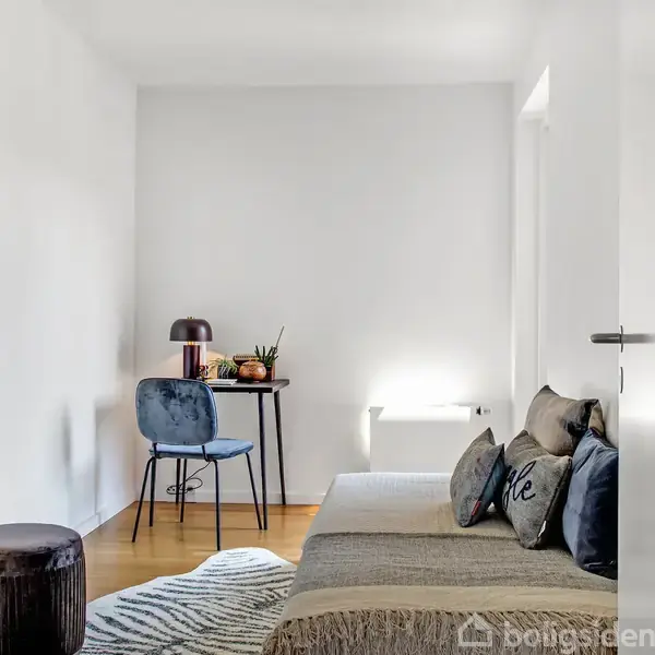 Et værelse med en sofa fyldt med puder står i forgrunden, mens et lille bord med en stol, lampe og planter ses i baggrunden. Lyse vægge og trægulve.