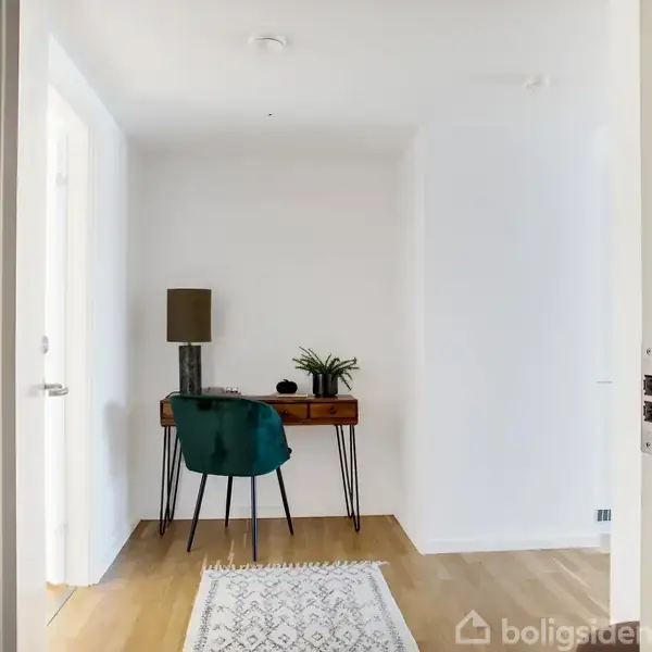 Et skrivebord med en lampe og en plante står i et lyst rum. En grøn stol står ved bordet på et trægulv. Rummet er minimalistisk indrettet.