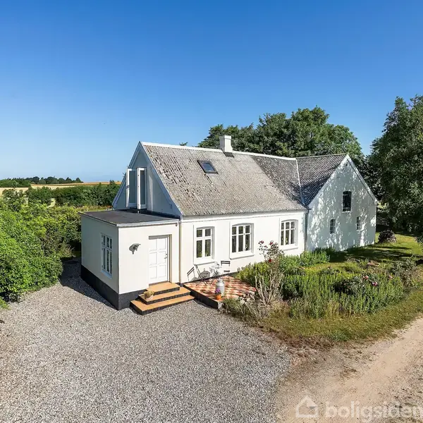 Hvidt landhus ligger ved grusvej, omgivet af grøn natur og træer. Huset har skråt tag, flere vinduer og en lille trappe ved indgangsdøren. Ingen synlig tekst i billedet.