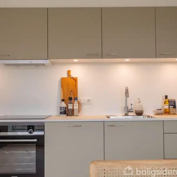 Køkkenskabe med beige fronter står over en bordplade med en vask og komfur. En vase med planter, skærebræt og flasker pynter. Lyse vægge giver et moderne look.