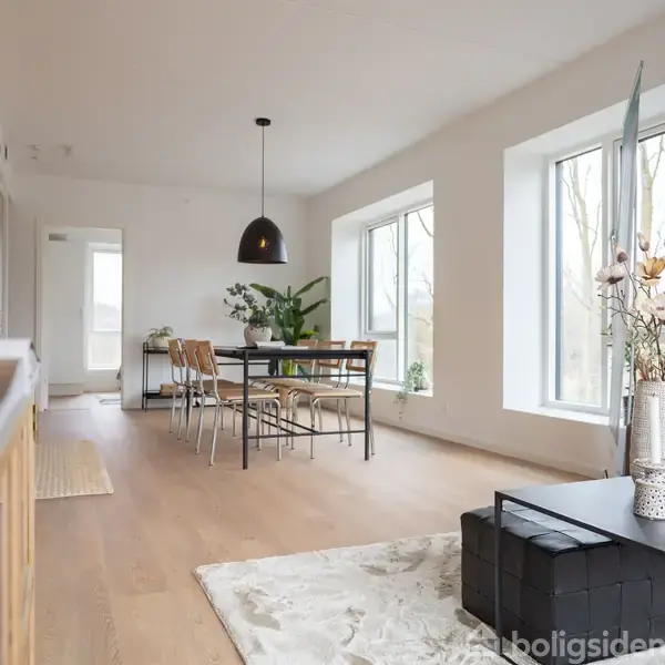 Stue med trægulv, sofabord og sofa til højre, spisebord med stole foran store vinduer. Hængelampe over bordet og plante i hjørnet skaber en lys og hyggelig atmosfære.