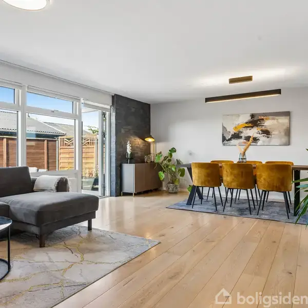 Sofa står stille i stuen med et lille bord ved siden af, omgivet af store vinduer og planter. Et spisebord med guldbelagte stole er i baggrunden under et kunstværk på væggen.
