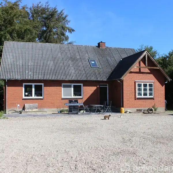 Rødt murstenshus med gråt tag står stille på en grusbelagt gårdsplads. Omringet af grønne træer under en klar blå himmel. En kat går på grusset. Ingen tekst til stede.