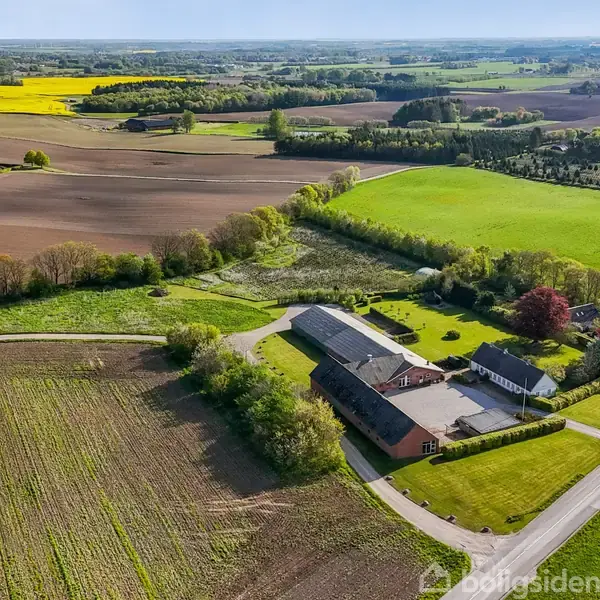 En landbrugsejendom med flere bygninger ligger omgivet af marker og træer. Veje fører til ejendommen gennem det åbne landskab, med farverige marker i baggrunden.