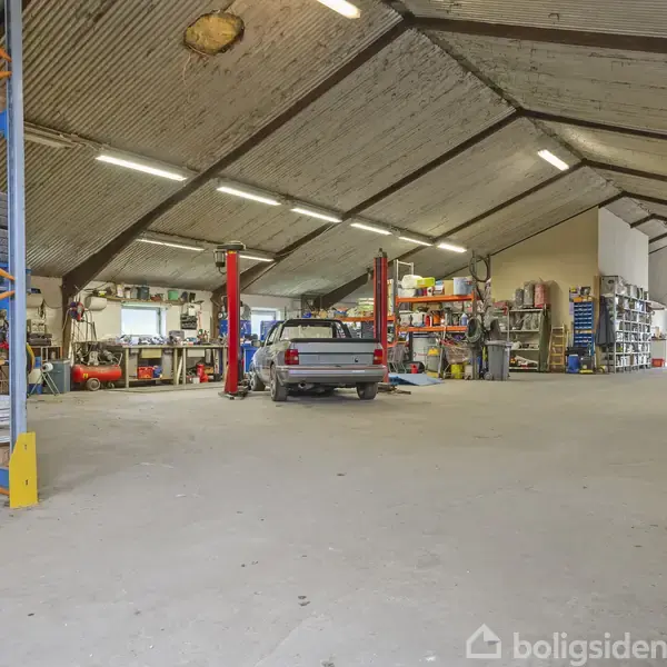 En bil er parkeret på en bilværkstedslift i en stor garage med skrå loftsbjælker. Reoler fyldt med dæk og værktøj står langs væggene i værkstedet.