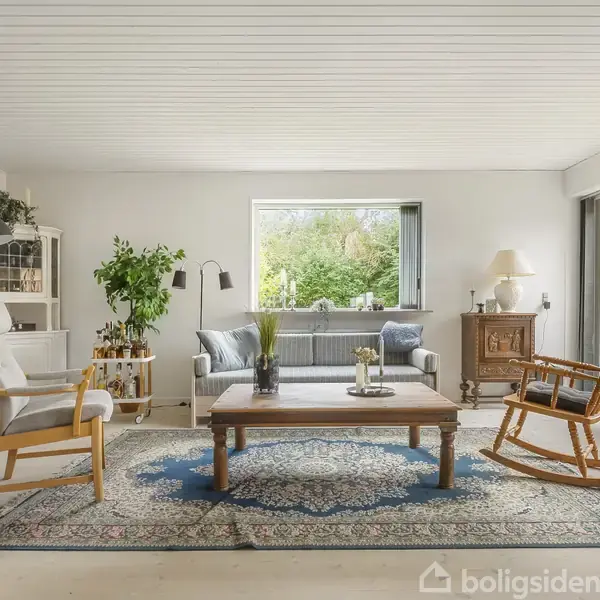 En lys stue har komfortable stole omkring et træbord på et orientalsk tæppe. Store vinduer giver udsigt til grønne omgivelser, mens en lampe og planter tilføjer hygge.