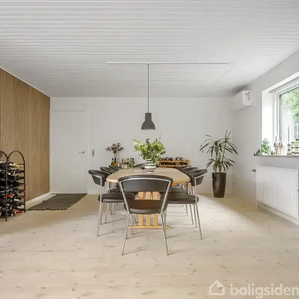 Spisebord med sorte stole står under en moderne lampe. Væggene er hvide med træpaneler. En vinreol og grønne planter pynter rummet. Store vinduer til venstre.