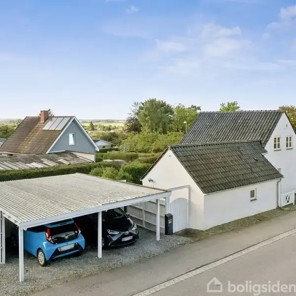 Et hvidt hus med skråt tag står ved siden af en carport med to biler, i et villakvarter med grønne træer og blå himmel.