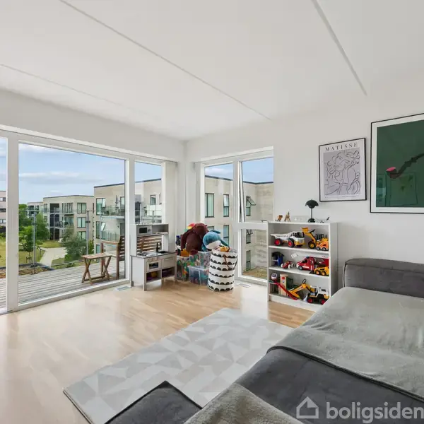 Et rummeligt værelse med en grå sofa og legetøjsreol. Store vinduer åbner mod en balkon med udsigt over en moderne boligblok. Trælegetøj er monteret på væggen.