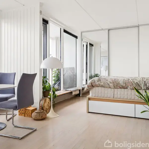 Et lyst rum indeholder en dobbeltseng med skydedøre bagved, en lille spisekrog med et rundt bord og to stole. En gulvlampe og flere planter dekorerer rummet.