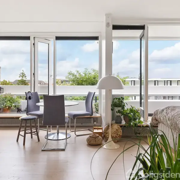 En lys stue med åbne glasdøre til altan, rundt spisebord med fire stole, omgivet af grønne planter og en sofa til højre. Store vinduer slipper lys ind.