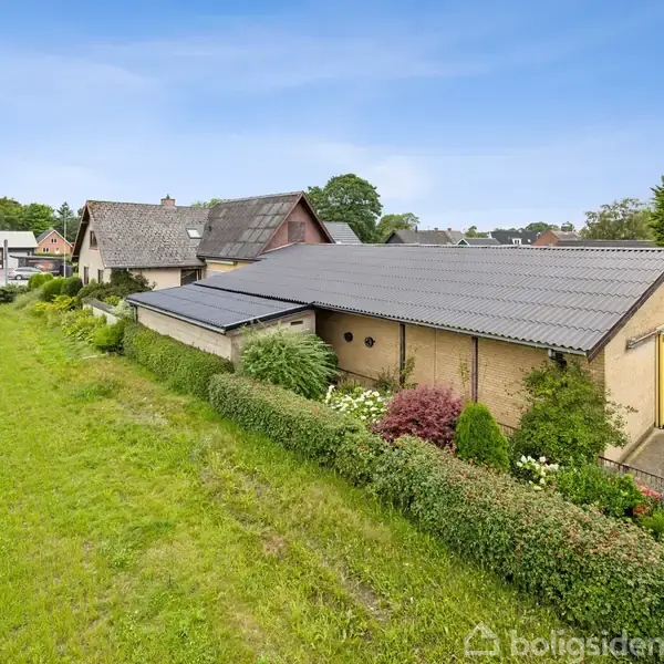 En gul bygning med gråt tag ligger i en grøn have med buske og blomster. Bagved er der flere huse og en blå himmel.