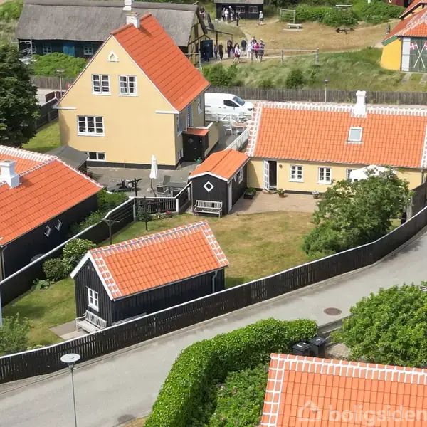 Flere gule huse med orange tage ligger i et tæt område omgivet af grønne buske og træer. En sti løber gennem beboelsen, som er indhegnet med lavt sort hegn.