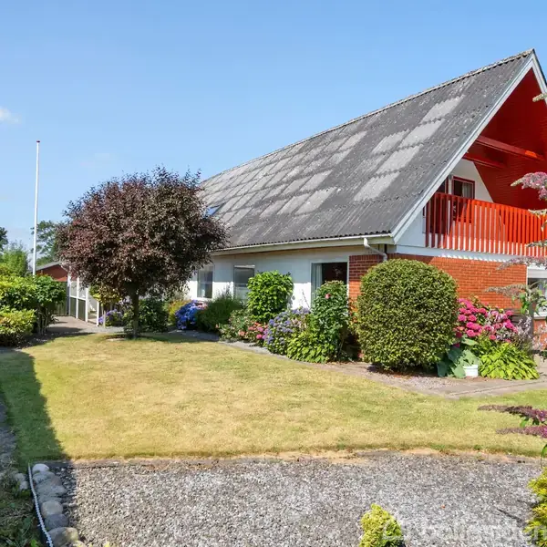 Et rødt, to-etagers hus med sadeltag står i en velplejet have omgivet af buske og blomster. Der er en grussti langs græsplænen og en træterrasse på første sal.