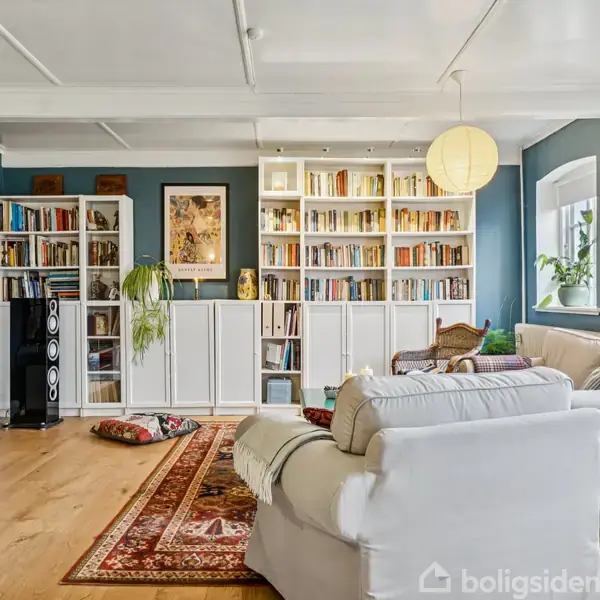 En stue med hvide sofaer står foran en stor reol fyldt med bøger. Et tæppe ligger på trægulvet, og et tv står i hjørnet. Blå vægge og store vinduer skaber rummet.