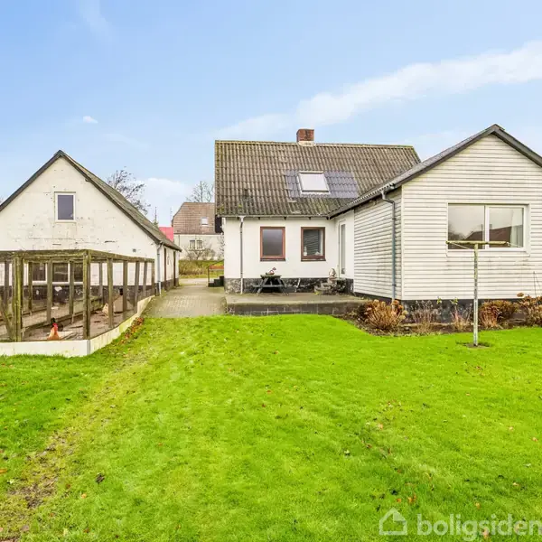 Hus står stille i en grøn have med et udhus til venstre. Sollys skinner på de hvide facader, omgivet af buske og træer i baggrunden.