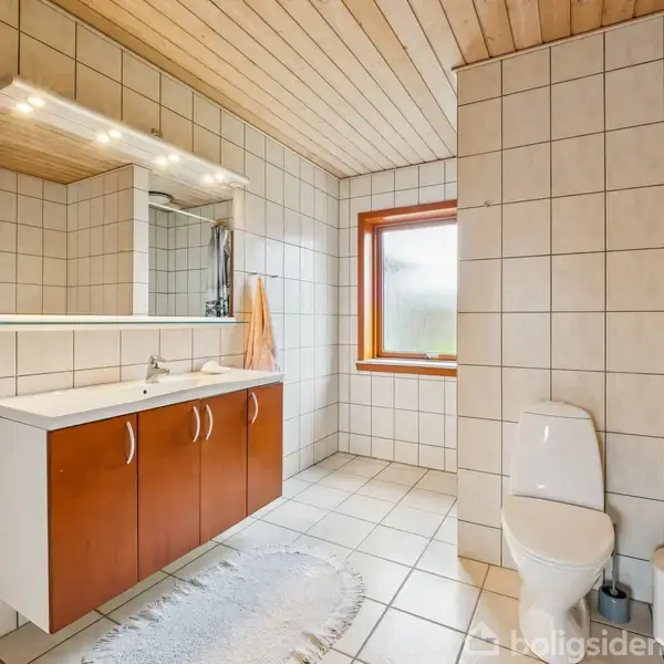 Badeværelse med hvide fliser, træloft og stort spejl. Brunt skab under vasken. Toilet står ved siden af en toiletpapirholder og to små beholdere. Vindue til højre giver naturligt lys.