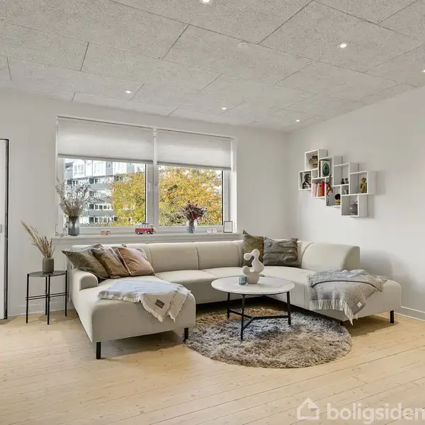 En lys stue med en hjørnesofa står i midten. Borde med dekorationer placeret foran sofaen. Store vinduer lader dagslys ind fra haven udenfor. Hylder hænger på væggen.