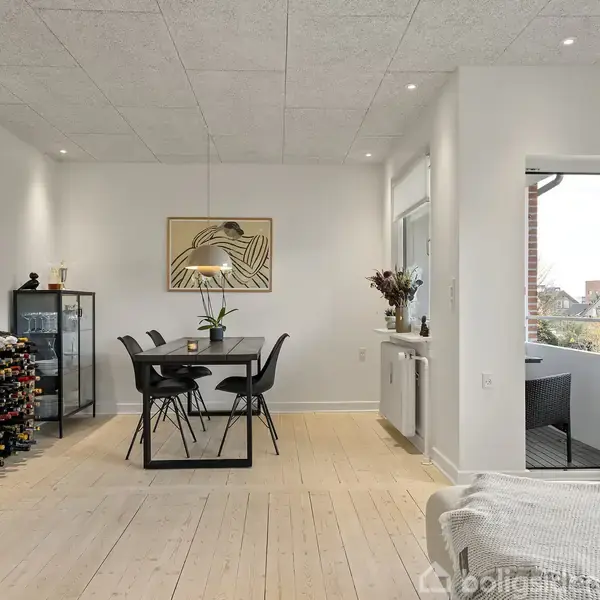 Spisebord med fire stole er centralt placeret i en lys, moderne spisestue. Et vinreol fyldt med flasker er til venstre, og et åbent vindue til højre.