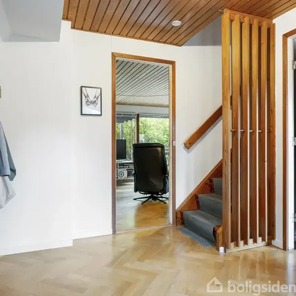 Entré med gulv af sildebensparket, trappe med trægelænder fører opad, jakker hænger til venstre. Dør fører til et værelse med kontorstol, set gennem døråbning.