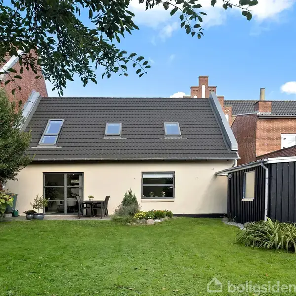 Et gult hus med gråt tag har en velplejet grøn have foran. Døre og vinduer viser ud til haven, omgivet af blomsterbede. Flere huse ses i baggrunden.