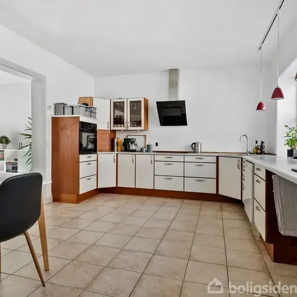 Et moderne køkken med hvide og træfarvede skabe, der danner en L-form rundt om arbejdsområdet. Røde lamper hænger over køkkenbordet. Et ur hænger på væggen. Et spisebord med sorte stole står i forgrunden.