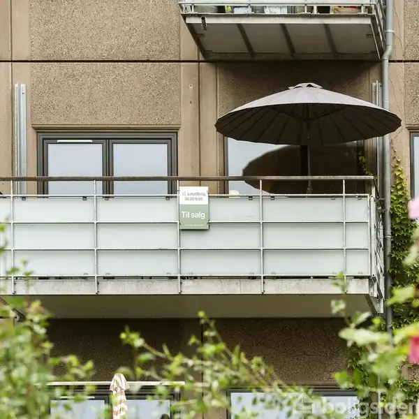 Balkon med en parasol står over en grøn have foran en murstensbygning. Et skilt på balkonen viser teksten "Lejlighed til salg".