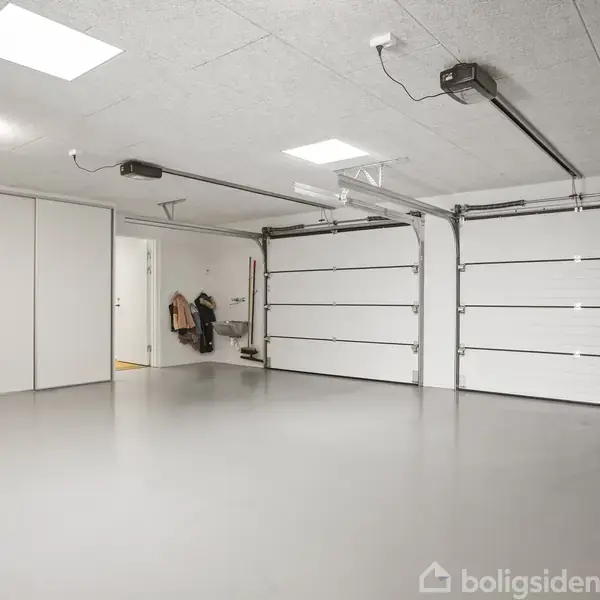 Garage med to hvide automatiserede porte, tom og rummelig. Vægge og gulv er lysegrå, og lysarmaturer hænger fra loftet. En dør i baggrunden fører til et andet rum.