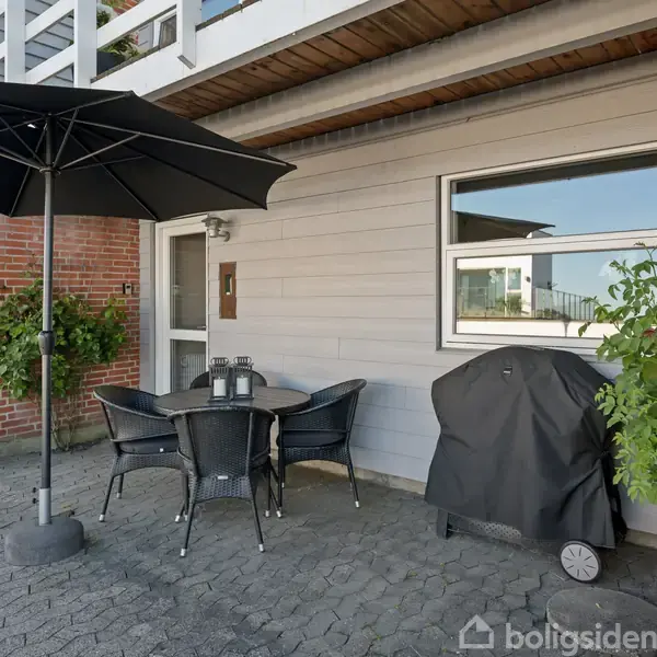 En terrasse med et rundt bord og fire sorte stole står under en stor parasol. En gasgrill dækket med presenning er placeret ved siden af. Omgivet af husfacader og planter.