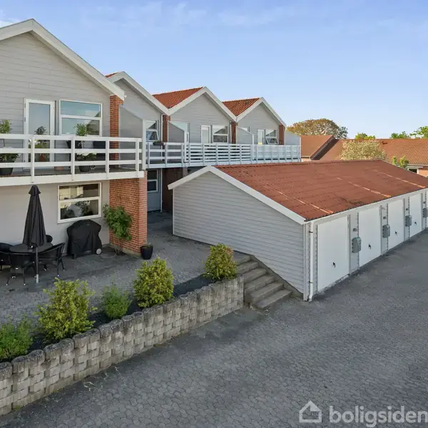 En række huse med røde tage og lyse facader står på en stenbelagt gårdsplads. I forgrunden ses en terrasse med bord, stole og planter. Parkering er til højre.